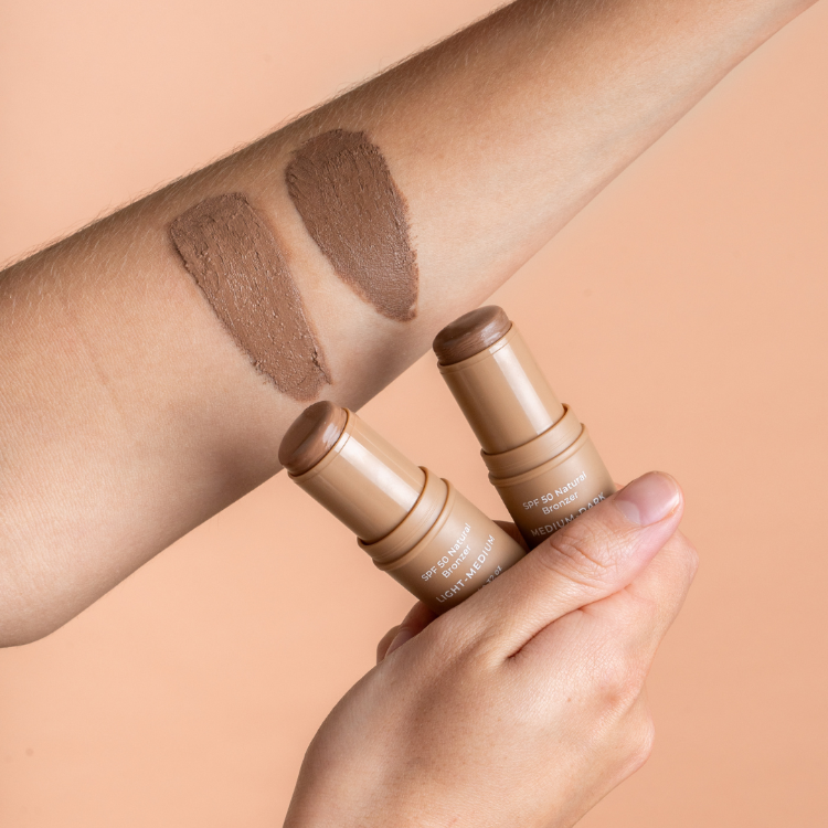 Two beige makeup sticks with swatches on a person's arm against a beige background