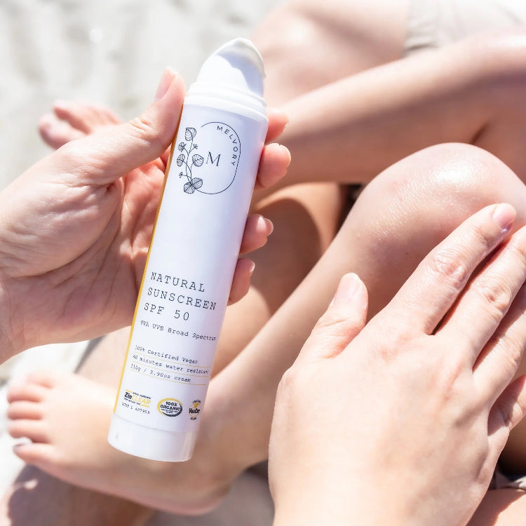 Hand holding a bottle of natural sunscreen with a child's legs in the background