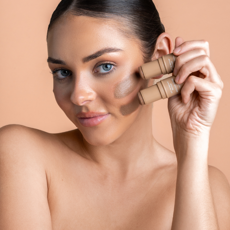 Woman applying makeup with a foundation stick on a beige background
